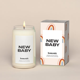 A lit New Baby Homesick candle displayed next to its boxed packaging on a dark cream background.