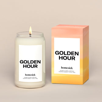 A lit Golden Hour Homesick candle displayed next to its boxed packaging on a dark cream background.