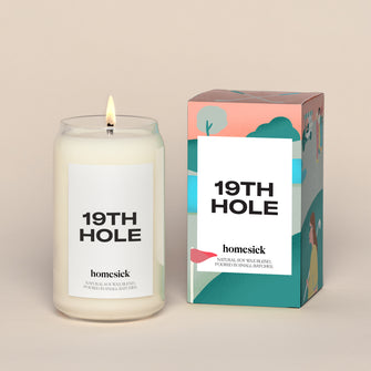 A lit 19th Hole Homesick candle displayed next to its boxed packaging on a dark cream background.