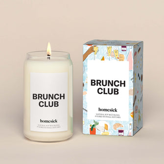 A lit Brunch Club Homesick candle displayed next to its boxed packaging on a dark cream background.