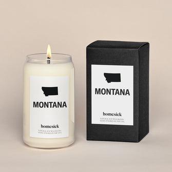 A lit Montana Homesick candle displayed next to its boxed packaging on a dark cream background.