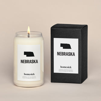 A lit Nebraska Homesick candle displayed next to its boxed packaging on a dark cream background.