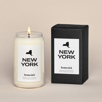 A lit New York City Homesick candle displayed next to its boxed packaging on a dark cream background.