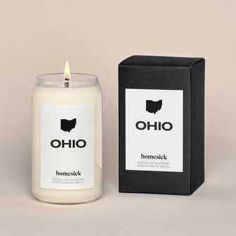 A lit Ohio Homesick candle displayed next to its boxed packaging on a dark cream background.