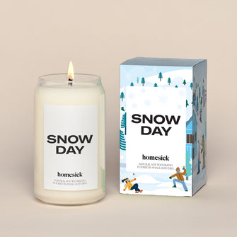 A lit Snow Day Homesick candle displayed next to its boxed packaging on a dark cream background.