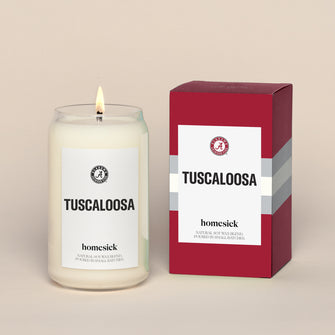 A lit Tuscaloosa Homesick candle displayed next to its boxed packaging on a dark cream background.