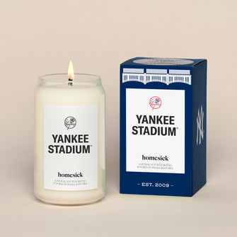 A lit Yankee Stadiu, Homesick candle displayed next to its boxed packaging on a dark cream background.